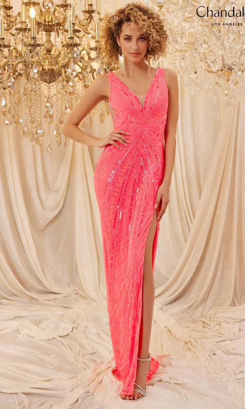 A woman in the Long Prom Dress 30091 by Chandalier, a sleeveless pink sequin v-neck formal gown with a thigh-high slit, stands before chandeliers and draped curtains.